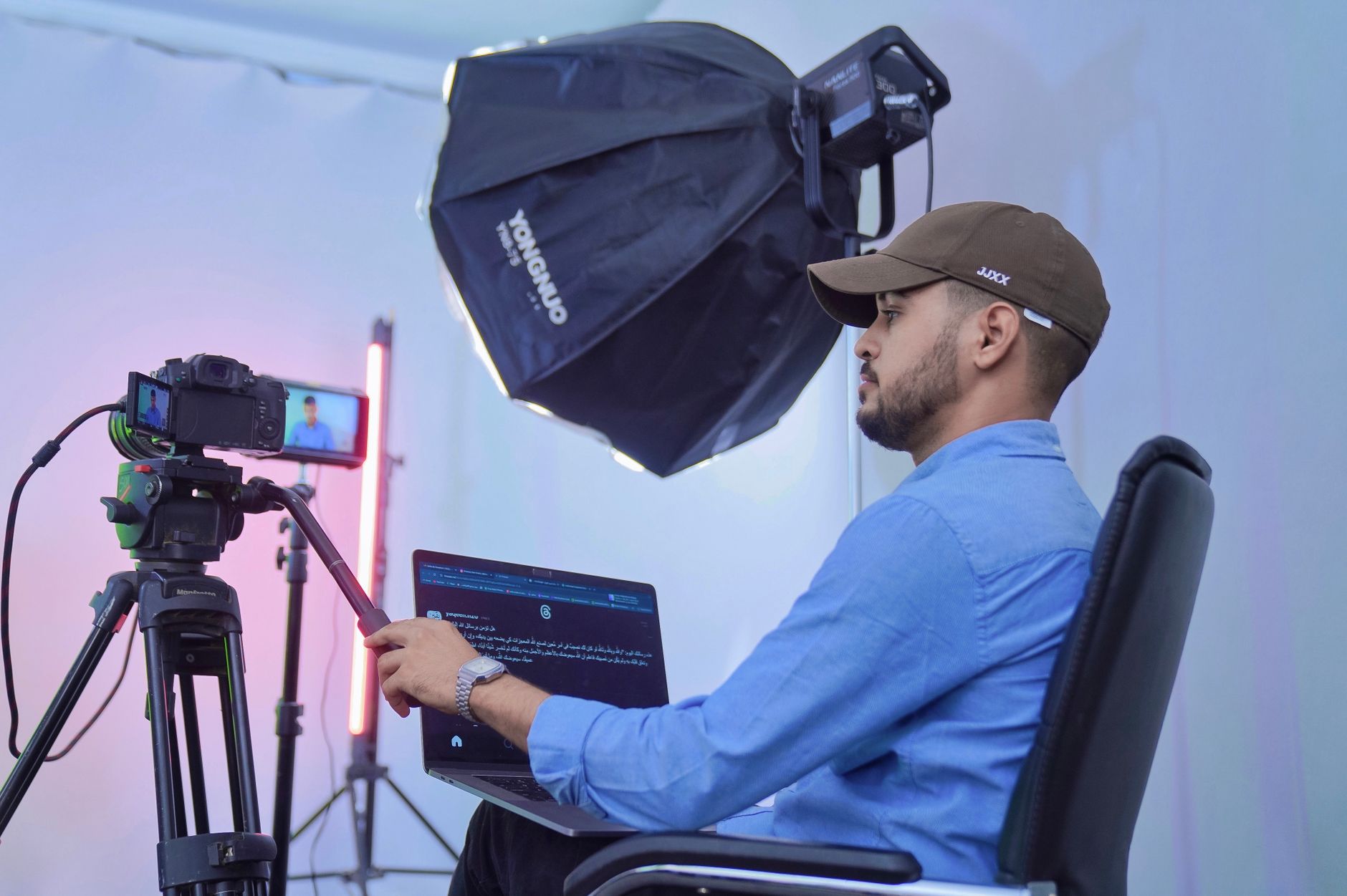 Un creador de contenido sentado junto a una cámara sobre trípode y un softbox, con un portátil sobre las rodillas, en un estudio improvisado
