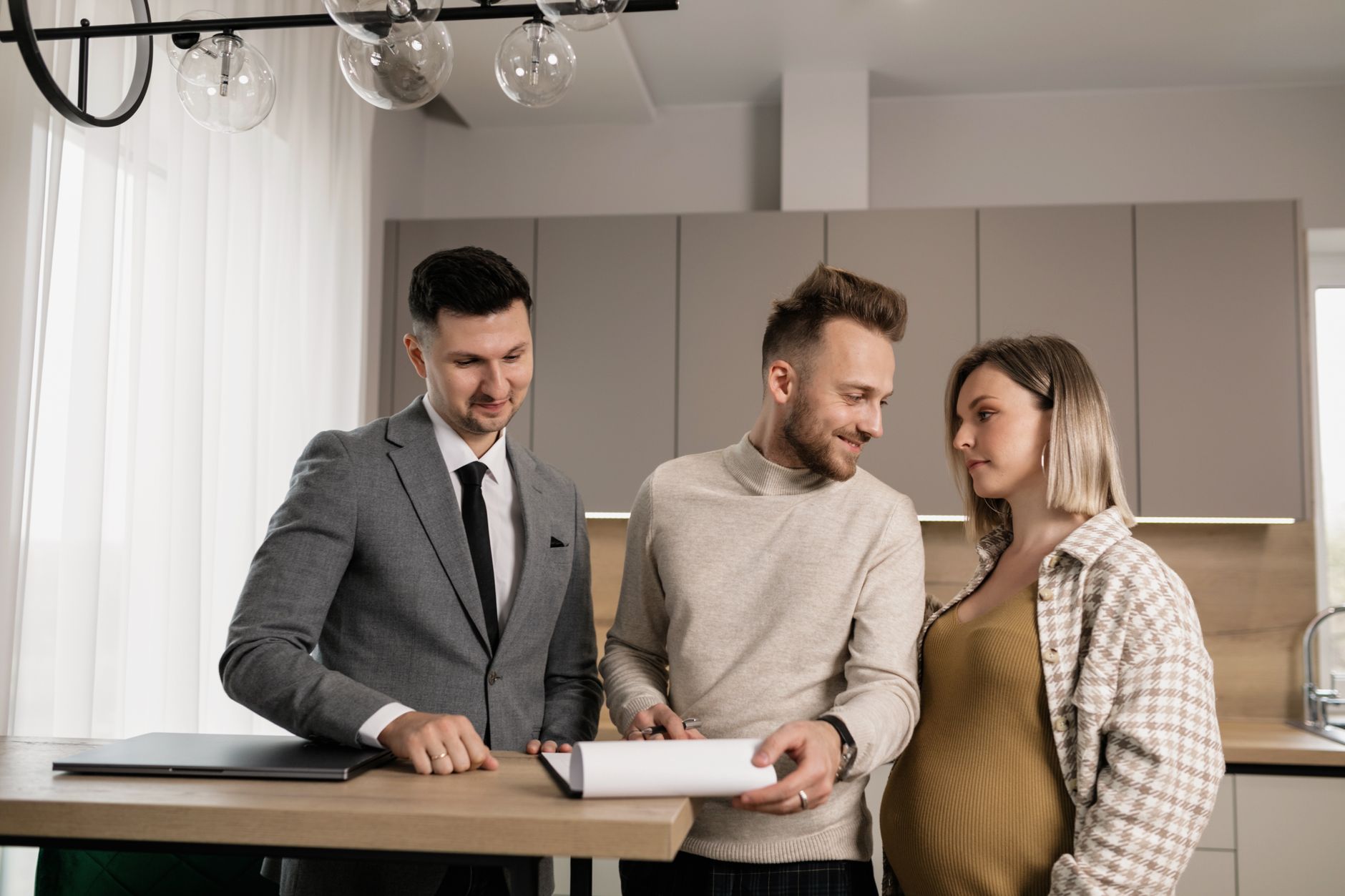 Una pareja y un agente inmobiliario con traje gris miran juntos un documento sobre una barra de cocina.