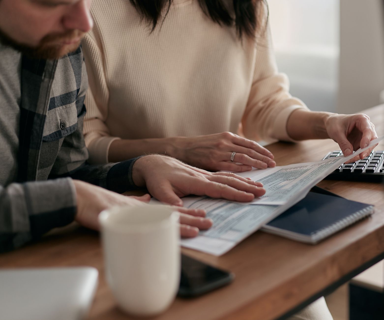 Una pareja repasa impresos y papeles sobre una mesa de madera, con una taza, un cuaderno y un teclado al lado.