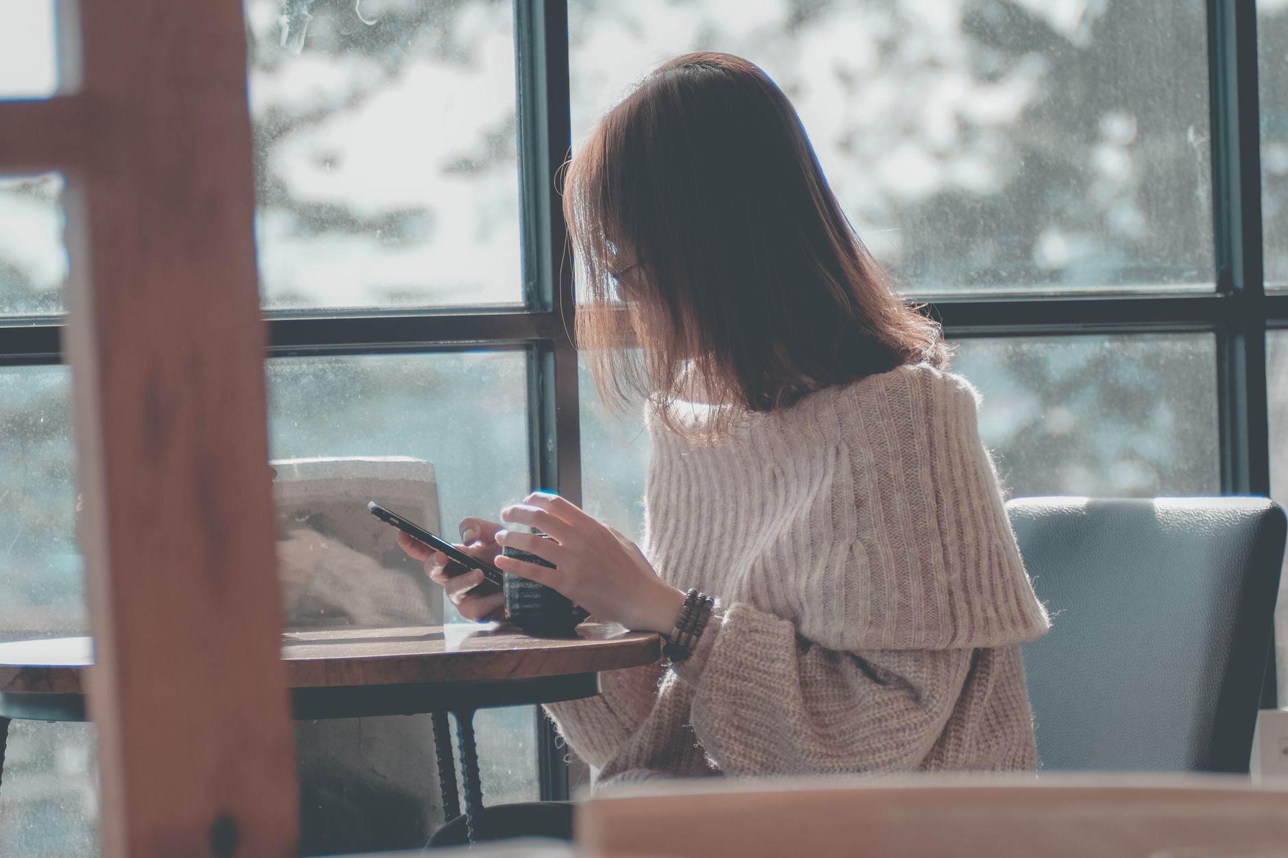 Mujer con un jersey de punto sentada junto a una ventana, con una taza de café y el móvil en la mano.