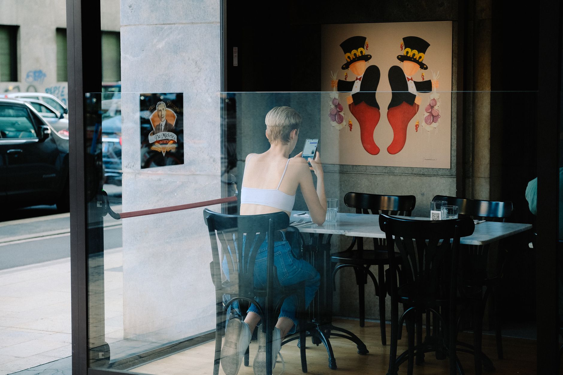Mujer sentada en una cafetería usando el móvil, vista a través del cristal del escaparate.
