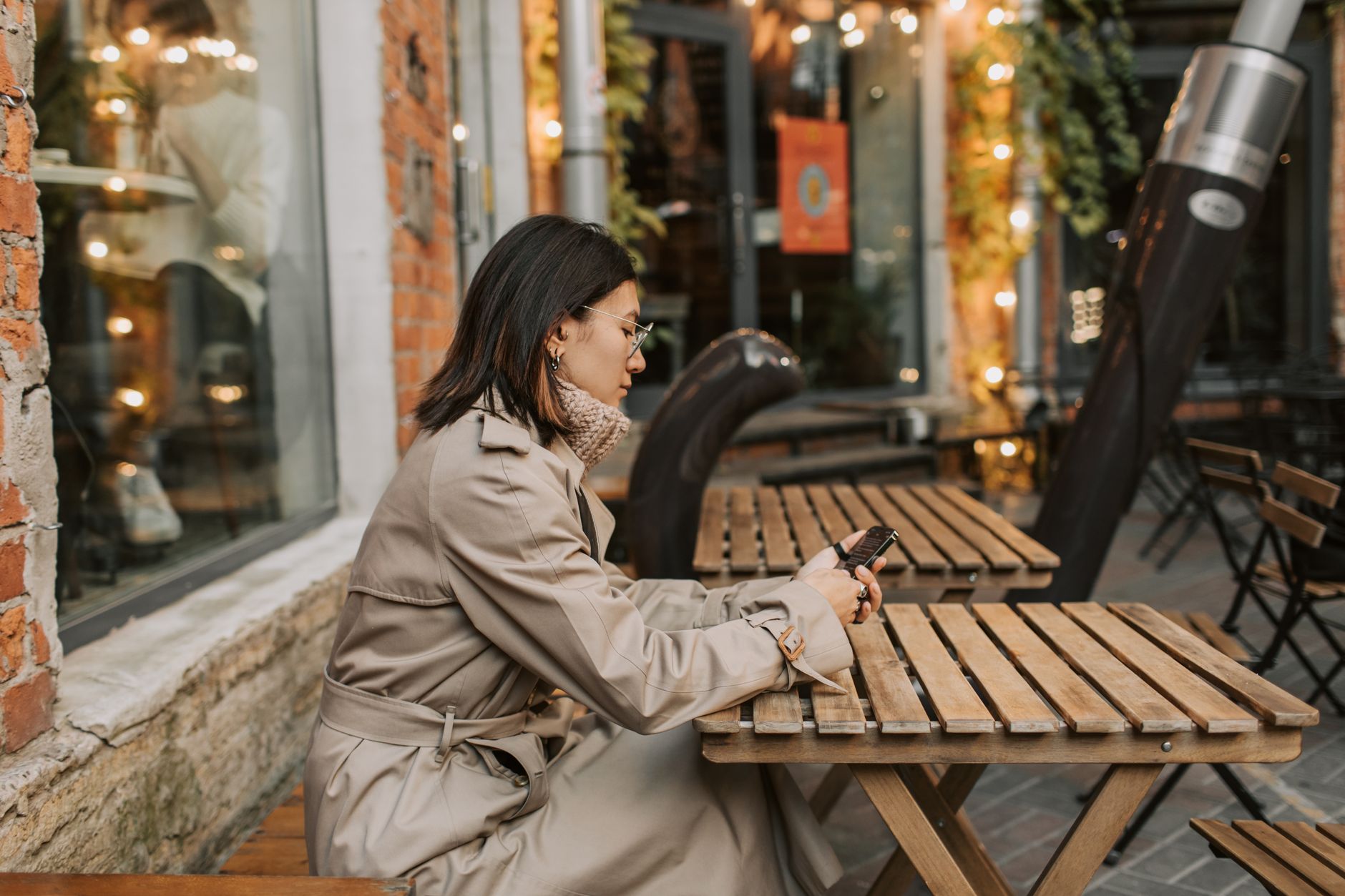 Mujer con gabardina beige y gafas mirando el móvil en la mesa de madera de una terraza.